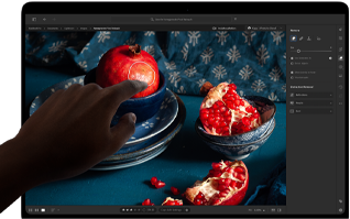 MacBook Pro con display PROLED XDR con l’immagine di una natura morta con melagrane e ceramiche e tessuti blu; una mano si avvicina allo schermo per interagire con l’immagine