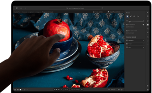 MacBook Pro con display PROLED XDR con l’immagine di una natura morta con melagrane e ceramiche e tessuti blu; una mano si avvicina allo schermo per interagire con l’immagine